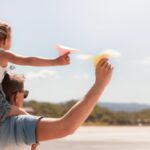 Homem com criança sobre os ombros com avião de papel na praia
