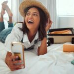 Mulher de chapéu deitada na cama com celular na mão e mala de viagem aberta