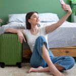 Mulher sentada no chão do quarto tirando selfie ao lado de mala de viagem